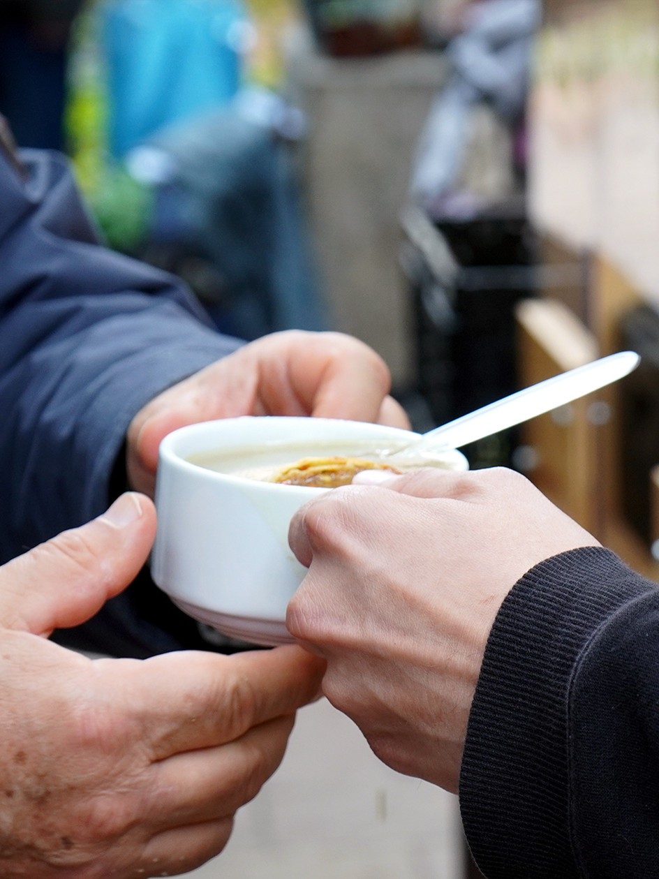 Ein Mitarbeiter im 's Häferl der Diakonie überreicht einem Gast draußen eine warme Suppe