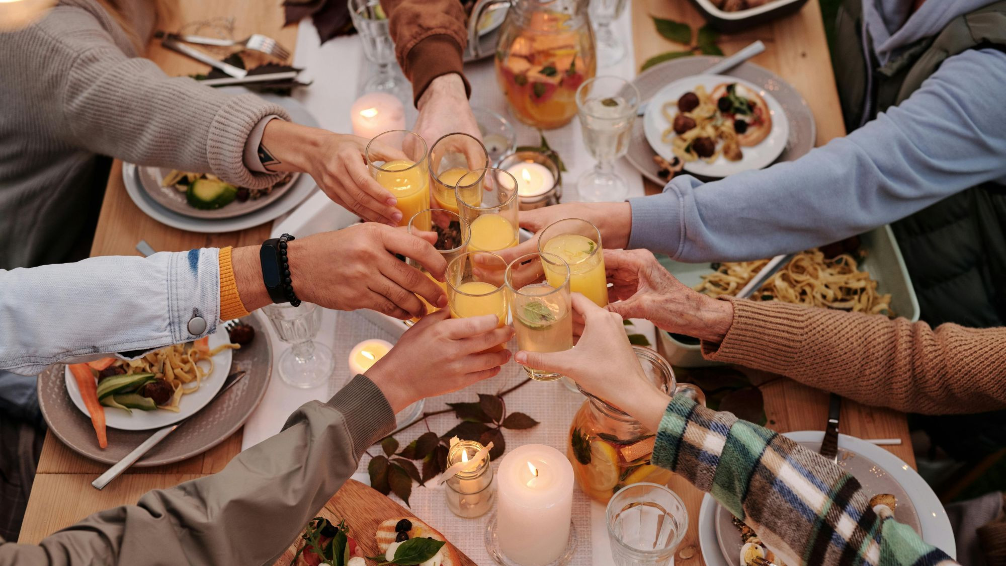 Ausgestreckte Arme mit Gläsern. Die Gläser werden zum Prost zusammengebracht. Ein Tisch mit Essen.