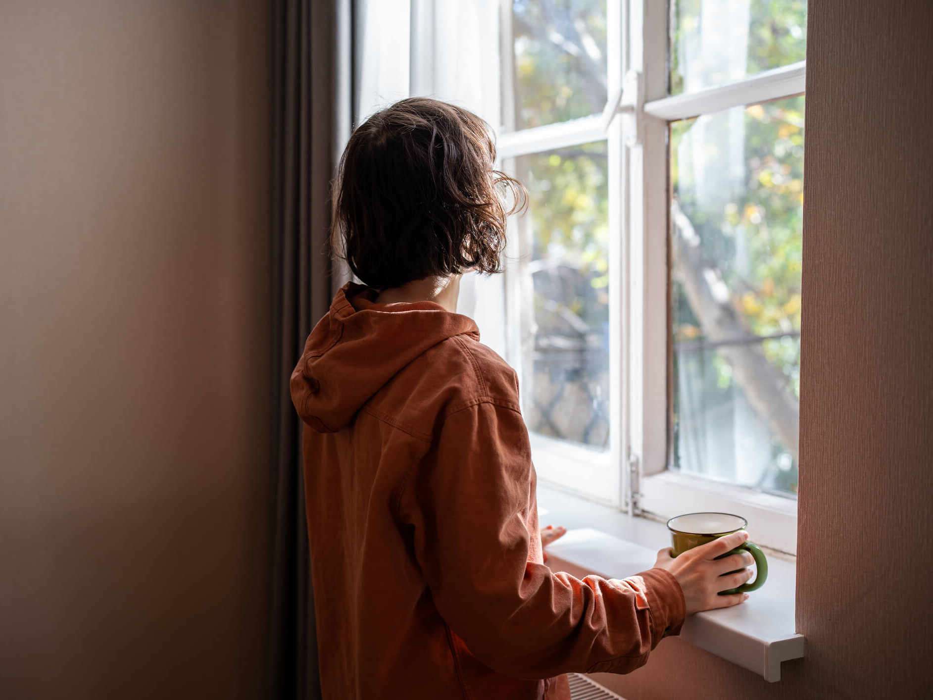 Die Frau steht am Fenster und blickt hinaus. In der Hand hält sie eine Tasse.