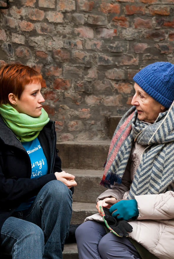 Eine junge Mitarbeiterin der Diakonie und eine ältere Frau sitzen auf einer steinernen Stiege, die ältere Frau erzählt, die jüngere hört zu