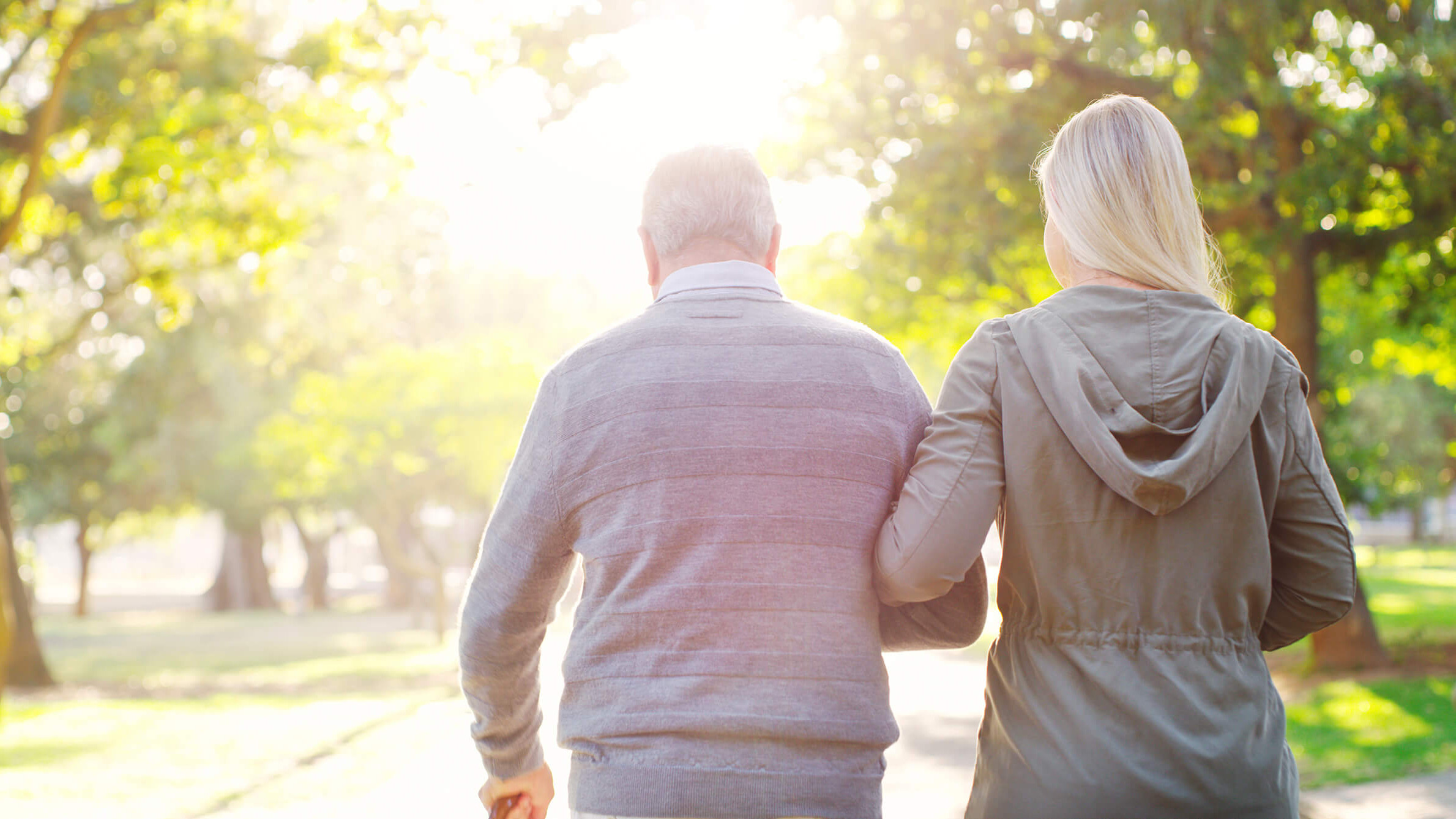 Ein alter Mann und eine jüngere Frau von hinten. Sie spazieren durch einen lichtdurchfluteten Park.