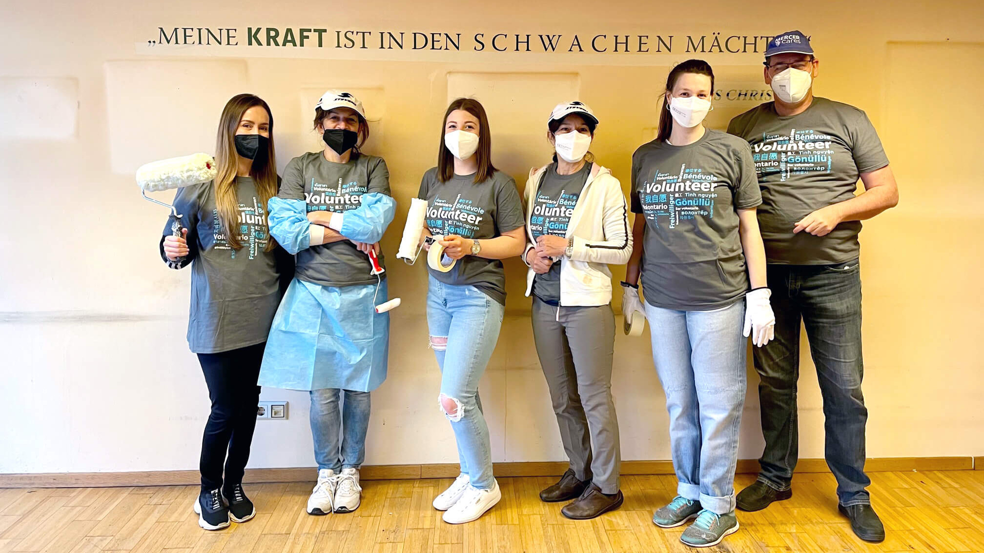 Eine Gruppe Menschen in Maleroutfits vor einer weißen Wand. Auf der Wand steht in schwarzer Schrift: Meine Kraft ist in den Schwachen mächtig.