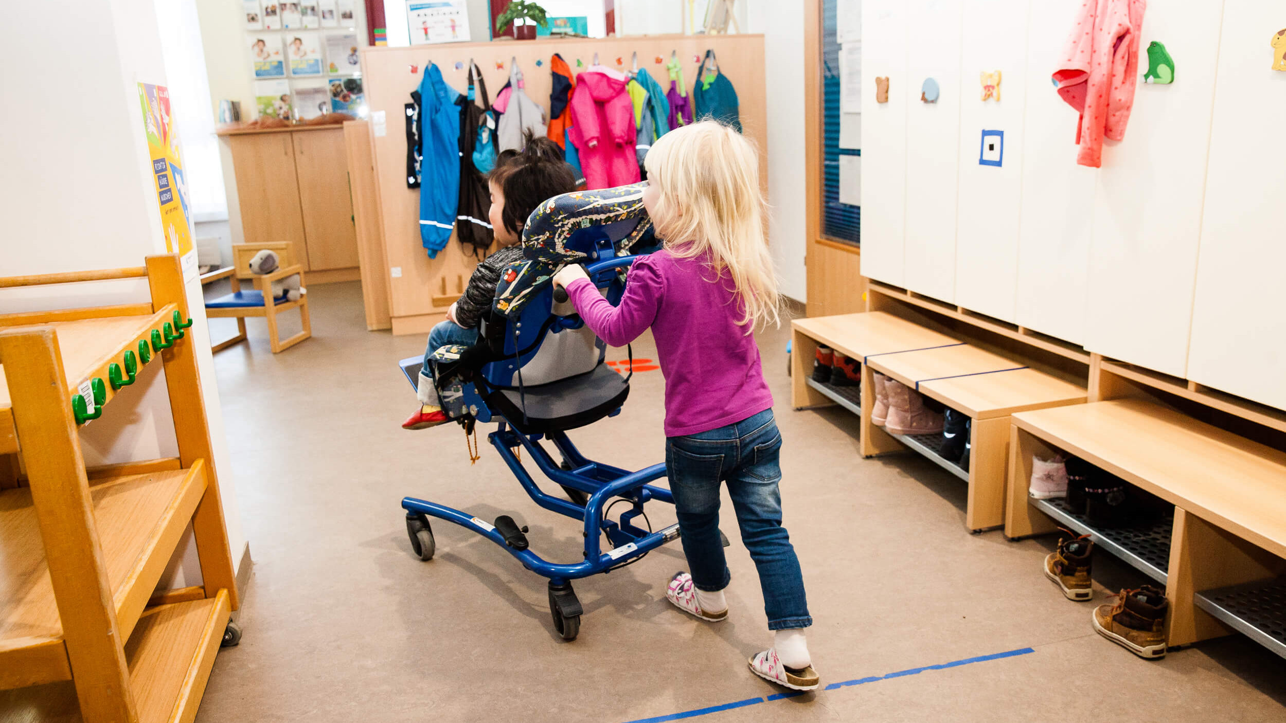 Ein Mädchen schiebt ein Mädchen im Rollstuhl durch einen Kindergarten.
