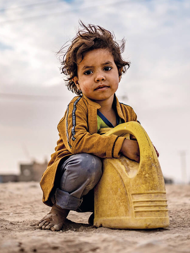 Ein Junge hockt am Boden, sieht in die Kamera, seine Hände umfassen einen Wasserkanister.